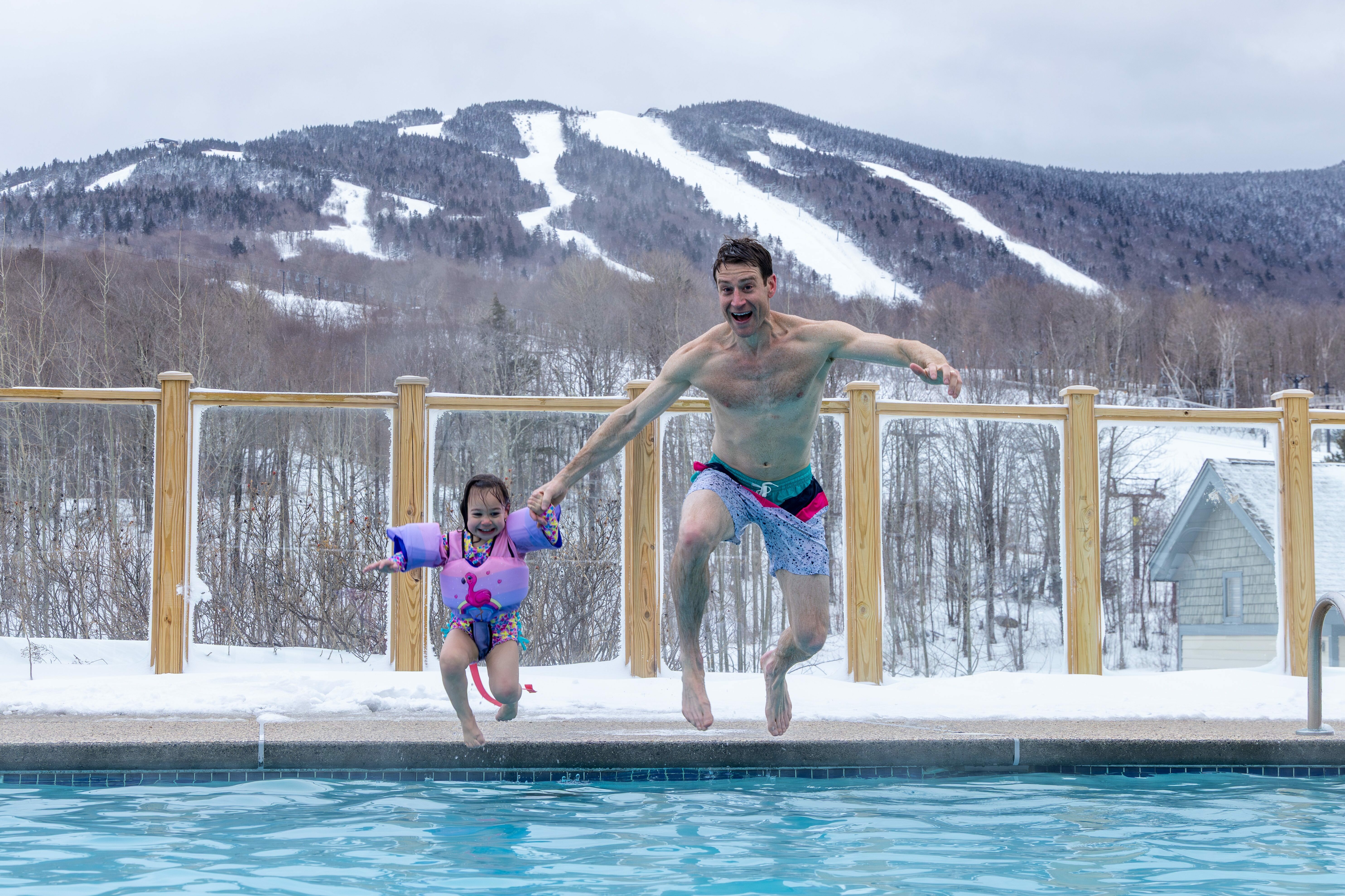A man and a girl jump into a pool surrounded by snowy mountains.