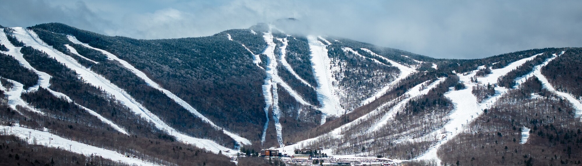Snow-covered ski slopes and trees under a cloudy sky in a mountainous landscape.