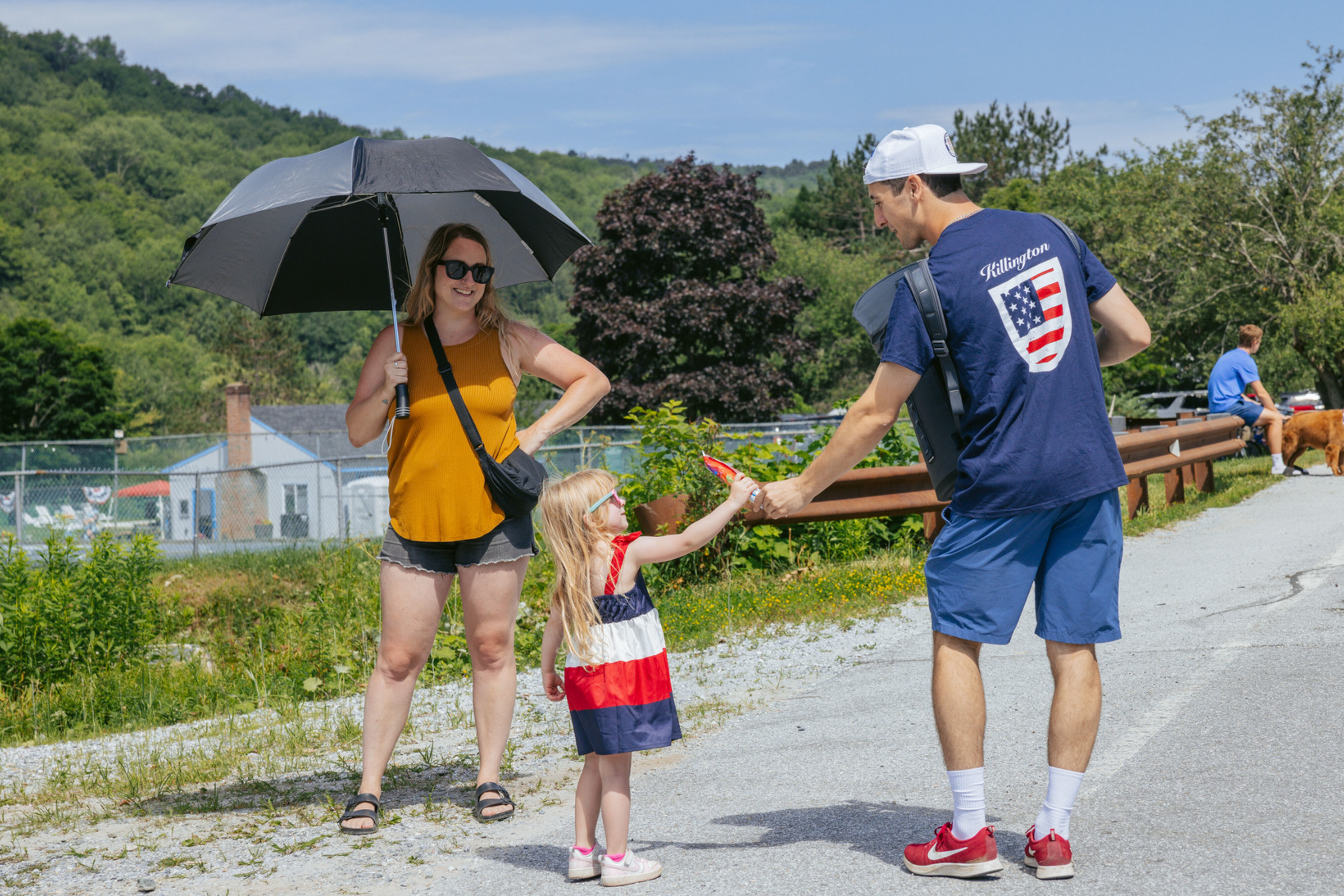 A woman holds an umbrella as a man hands a treat to a young girl in a patriotic dress.