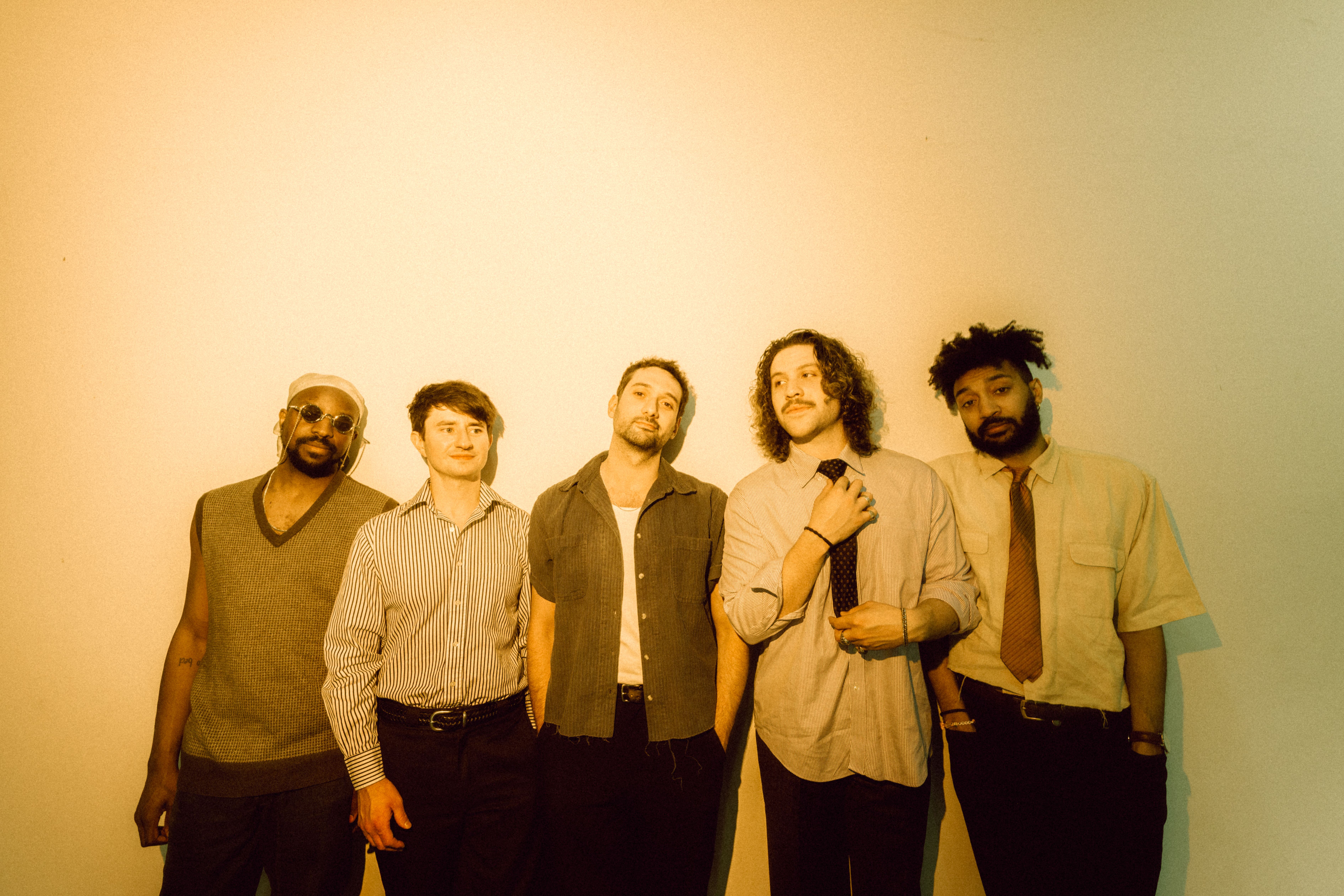 Five men in stylish outfits pose together against a warm-toned backdrop.