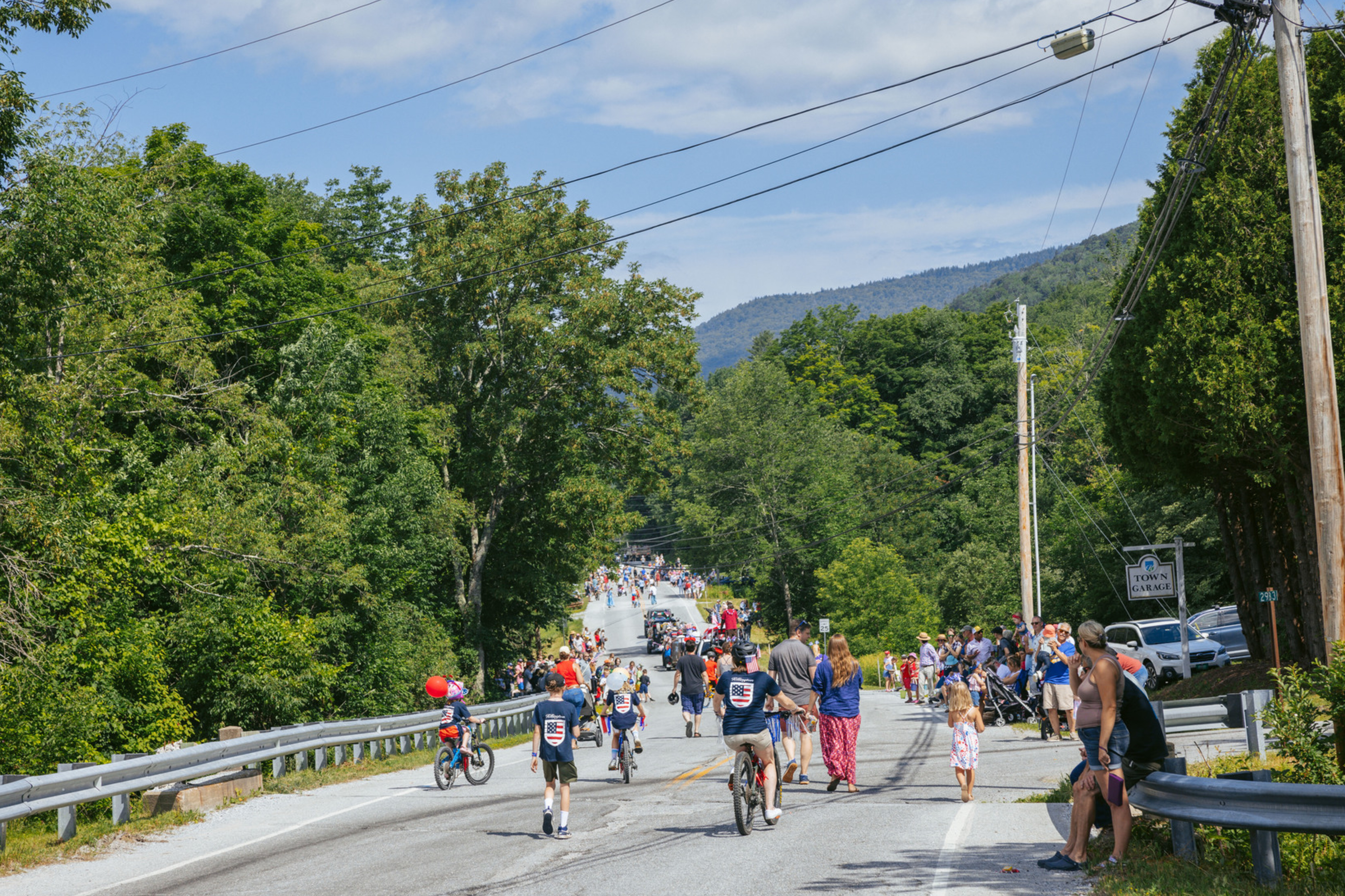A festive street scene with families walking and biking along a tree-lined road.