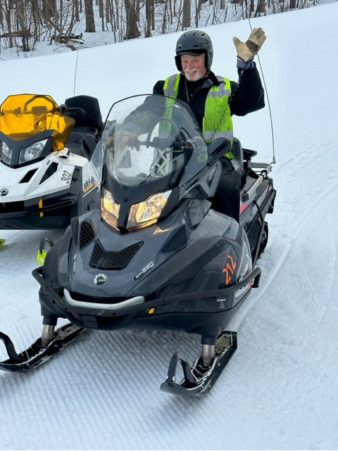 A smiling person waves while seated on a snowmobile in a snowy area.