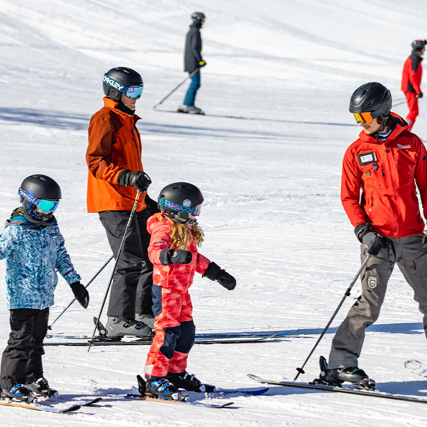 A ski instructor assists children on the slopes, surrounded by skiers in a snowy landscape.