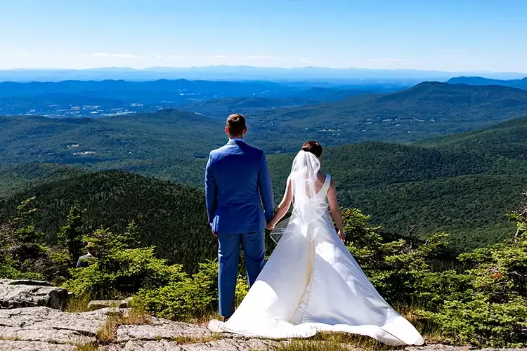Bride and groom holding hands, gazing at scenic mountain view.