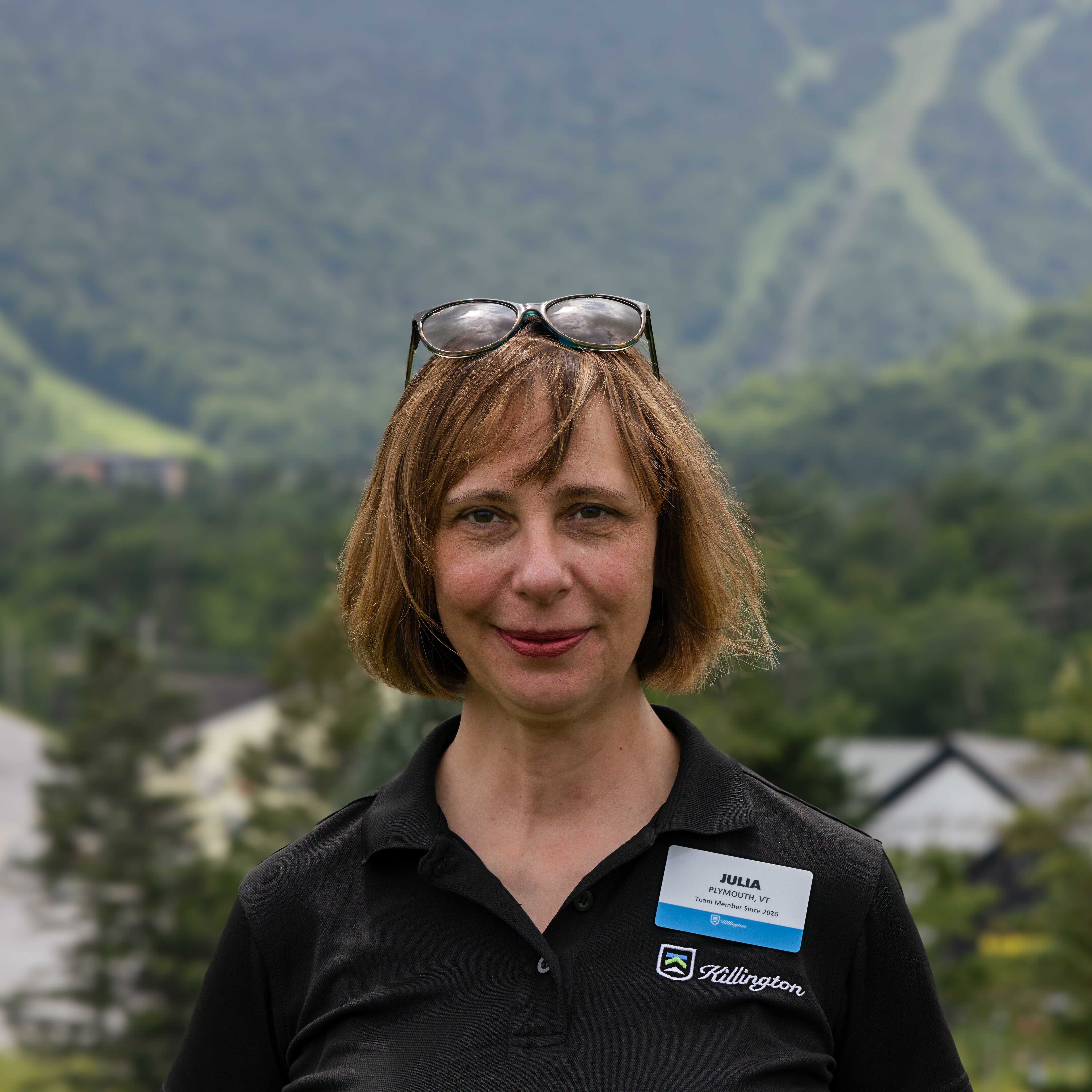 Woman with short hair and glasses stands outdoors with mountains in the background.