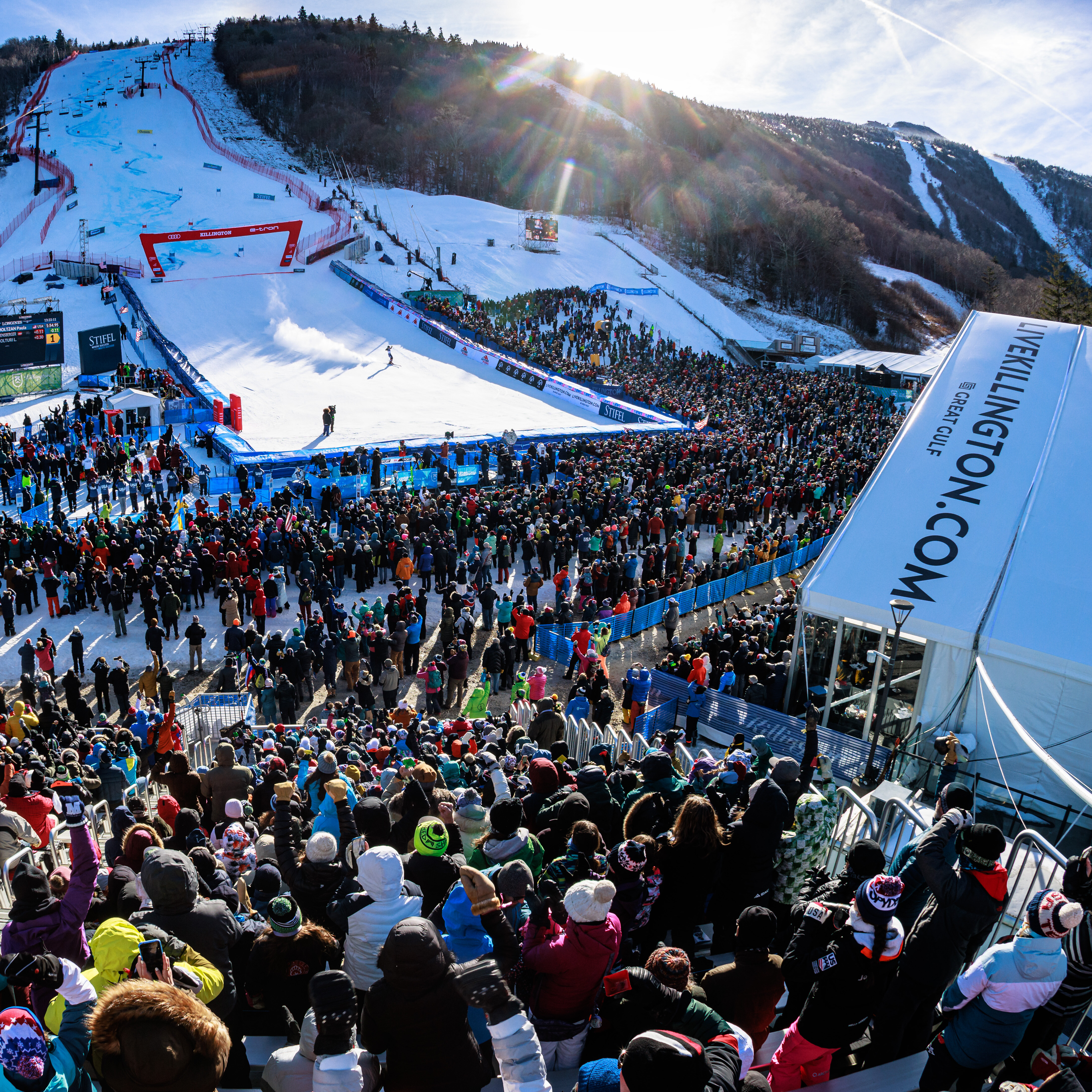 Crowd gathered at a snowy ski event with slopes and mountains in the background.