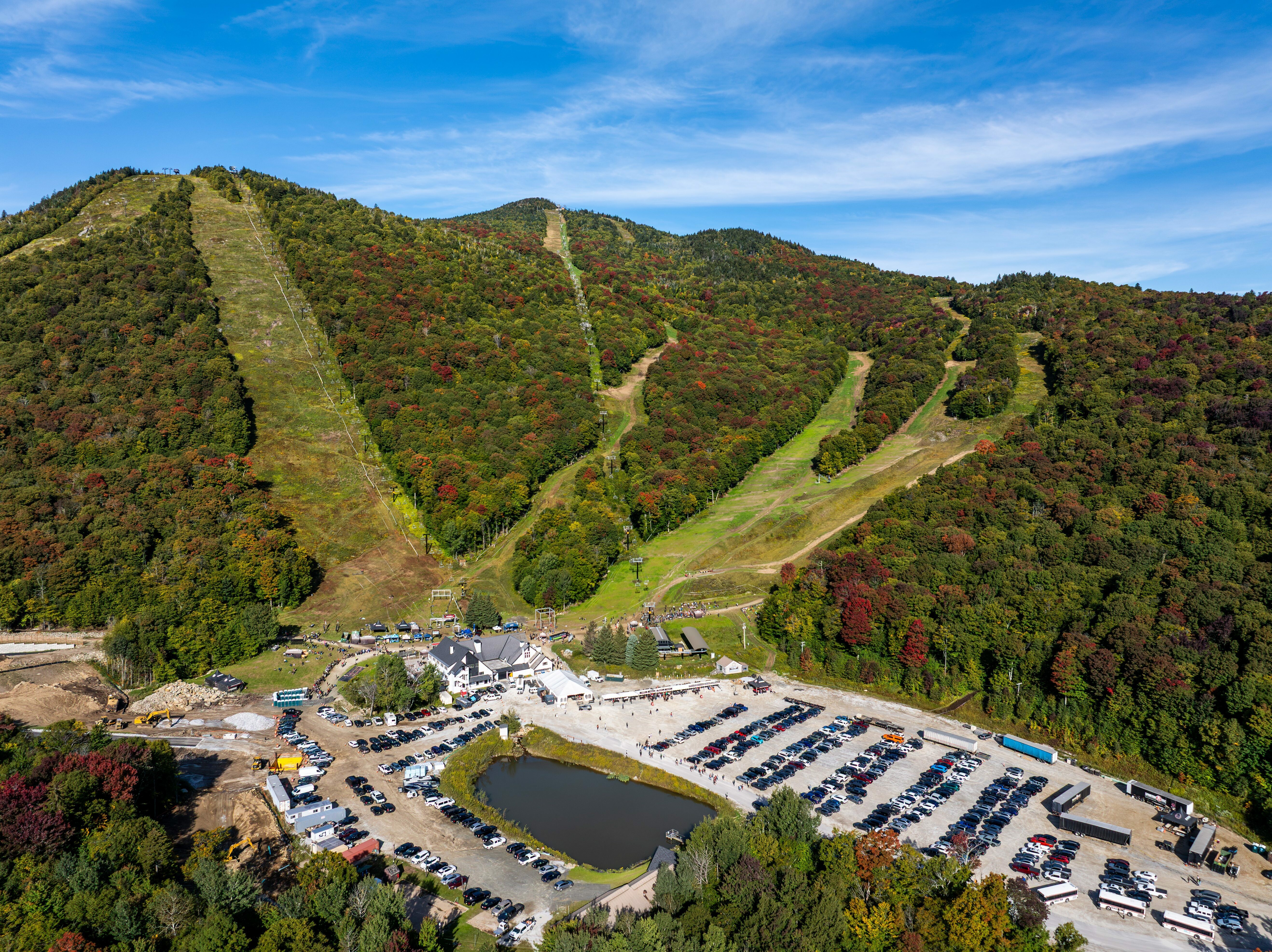 Aerial view of fall-colored hills, a ski area, and a busy parking lot near a pond.