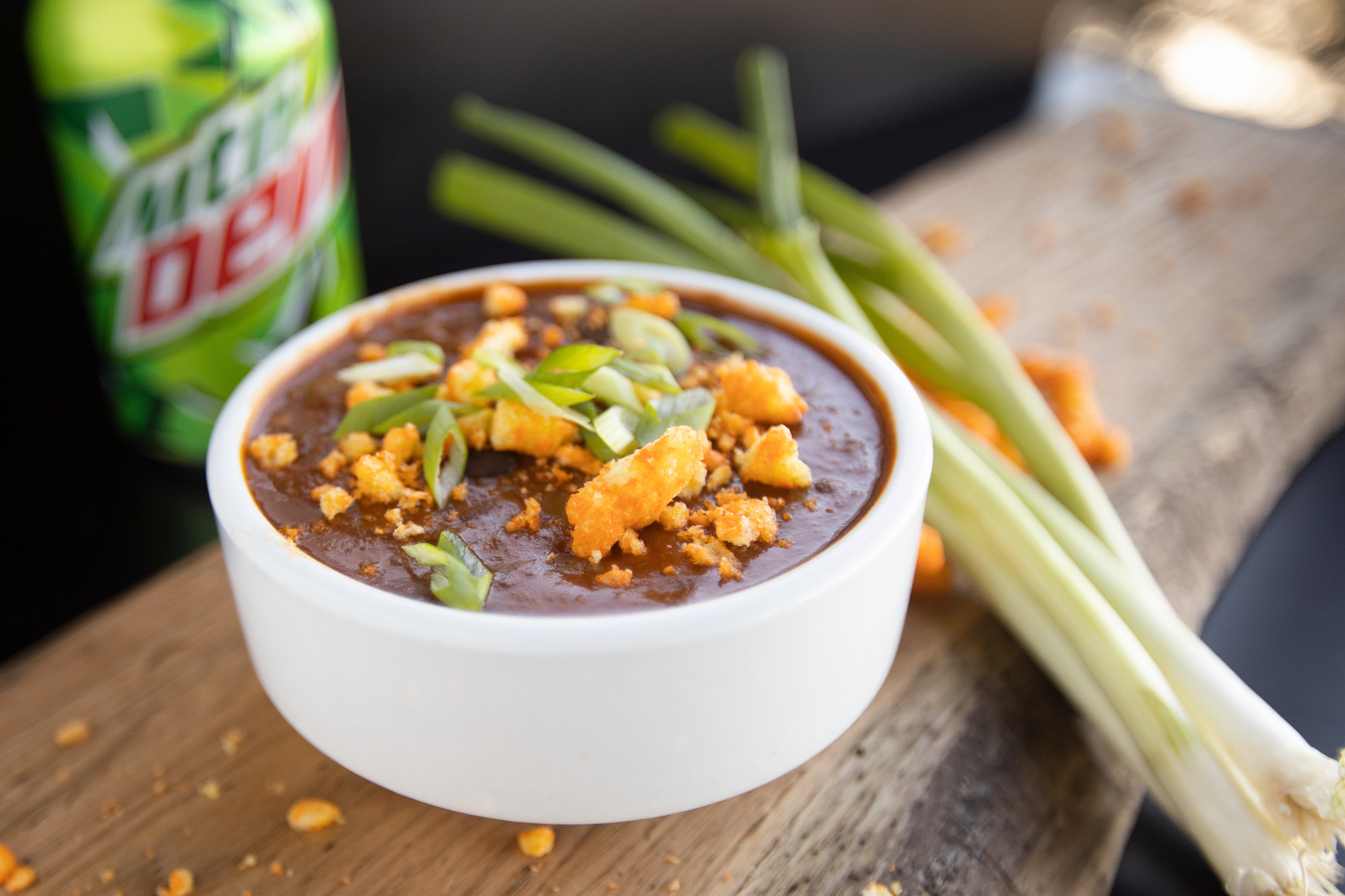 Bowl of chili topped with green onions and cornbread on a wooden board, with a Mountain Dew can.
