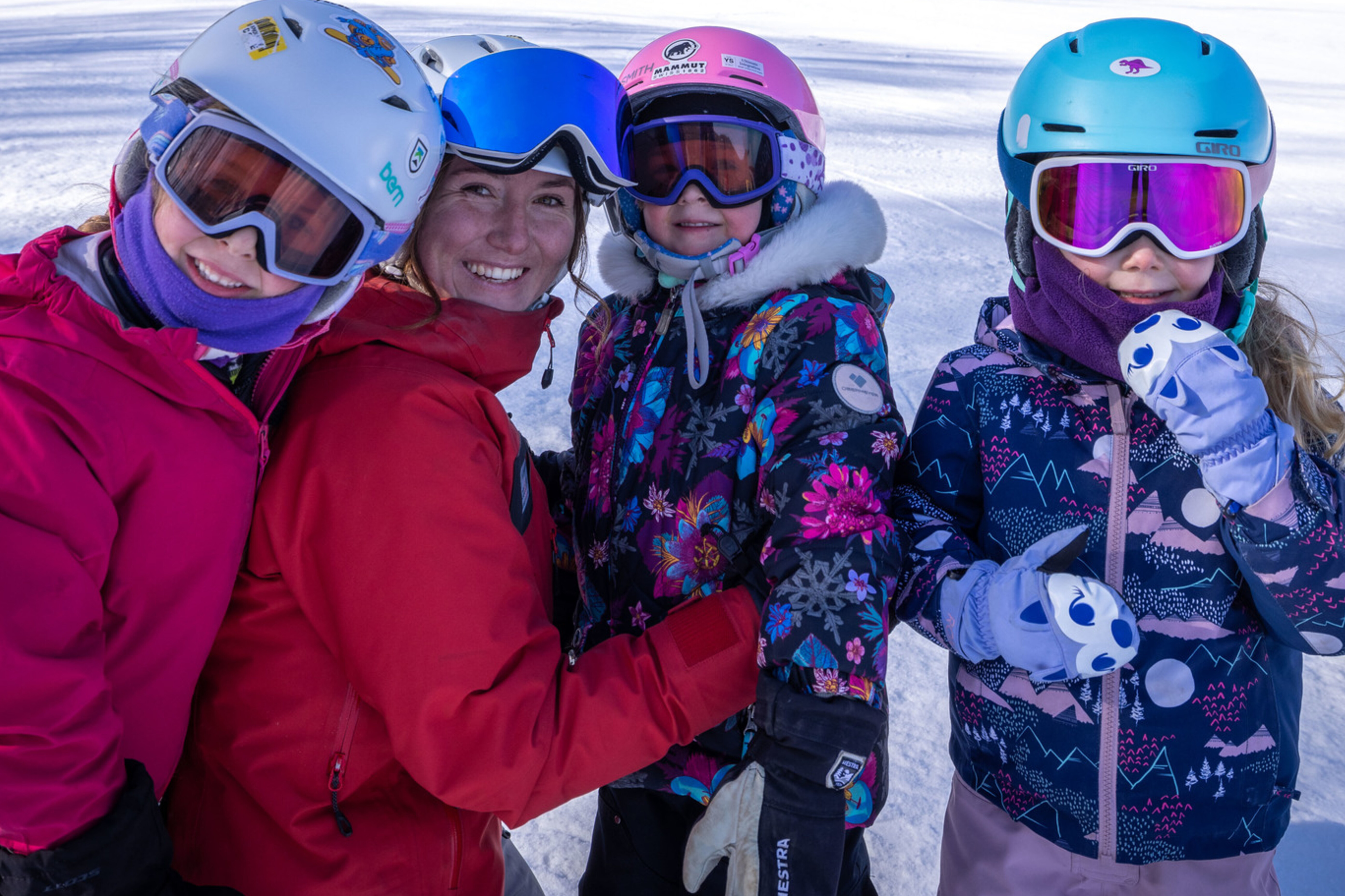 A smiling woman poses with three children in ski gear on a snowy slope.