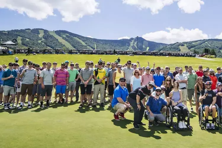 A large group of people poses together on a golf course with mountains in the background.