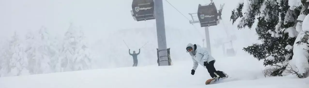 A snowboarder rides through deep snow with a ski lift in the background.