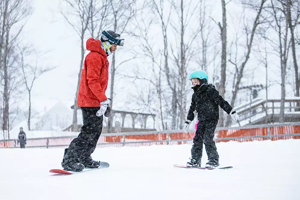 A child snowboards with an instructor in a snowy landscape.
