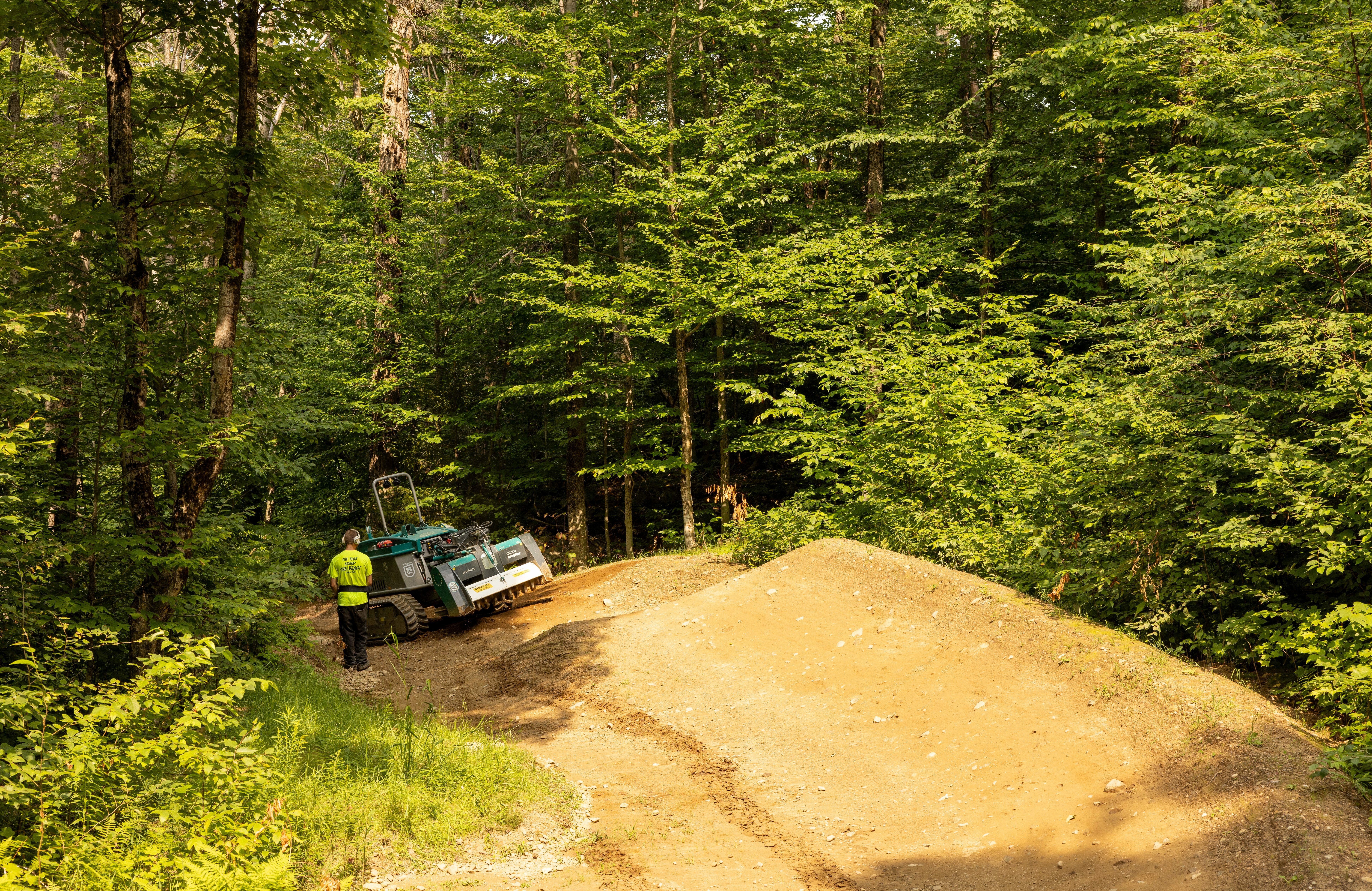 A worker stands beside a machine on a mountain bike trail in a lush green forest.