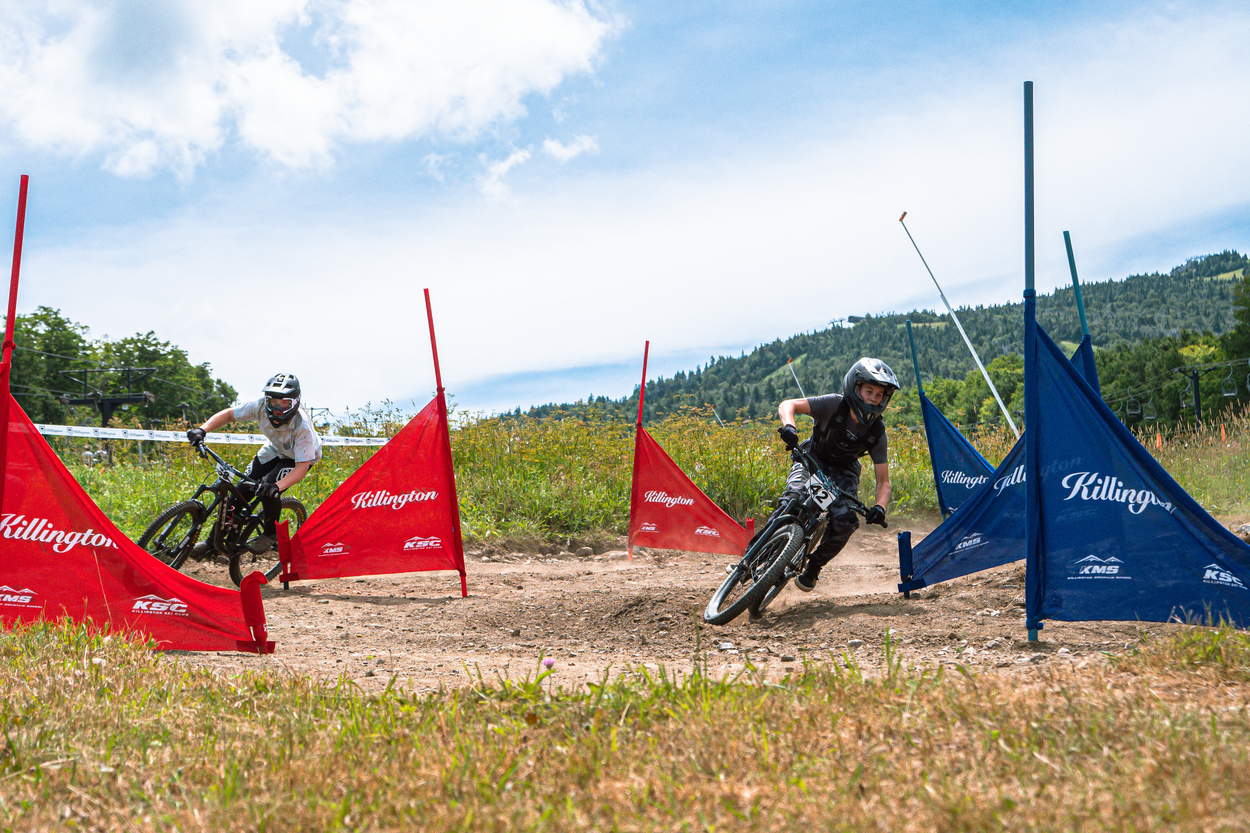 Two mountain bikers race through red and blue flags on a dirt track with hills in the background.