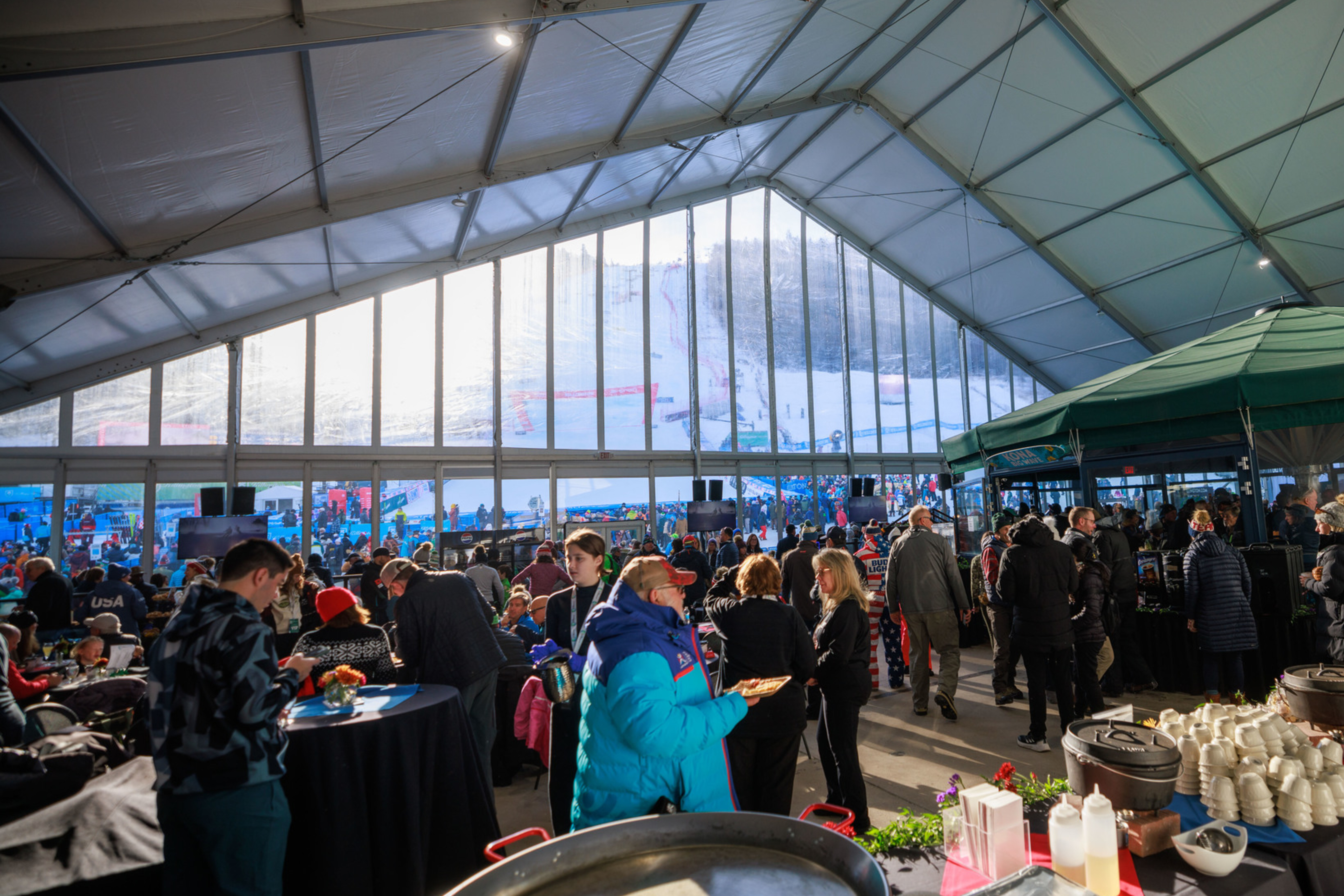 Busy indoor event with people mingling, food stations, and a view of a snowy slope outside.