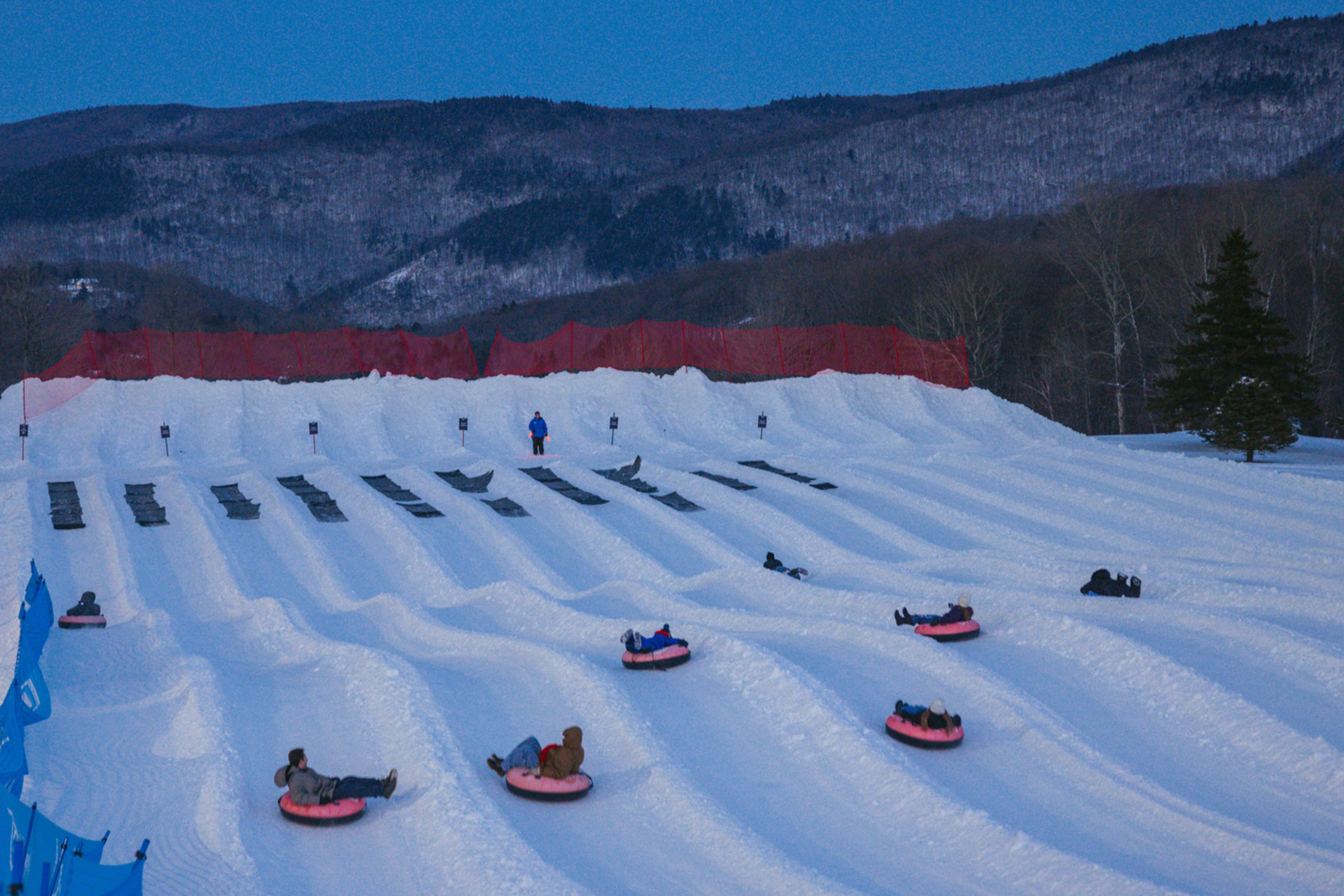 Snow tubing hill with several people on tubes and a snowy landscape at dusk.