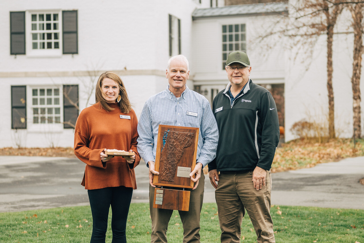 Three people stand outdoors, holding an award in front of a white building.