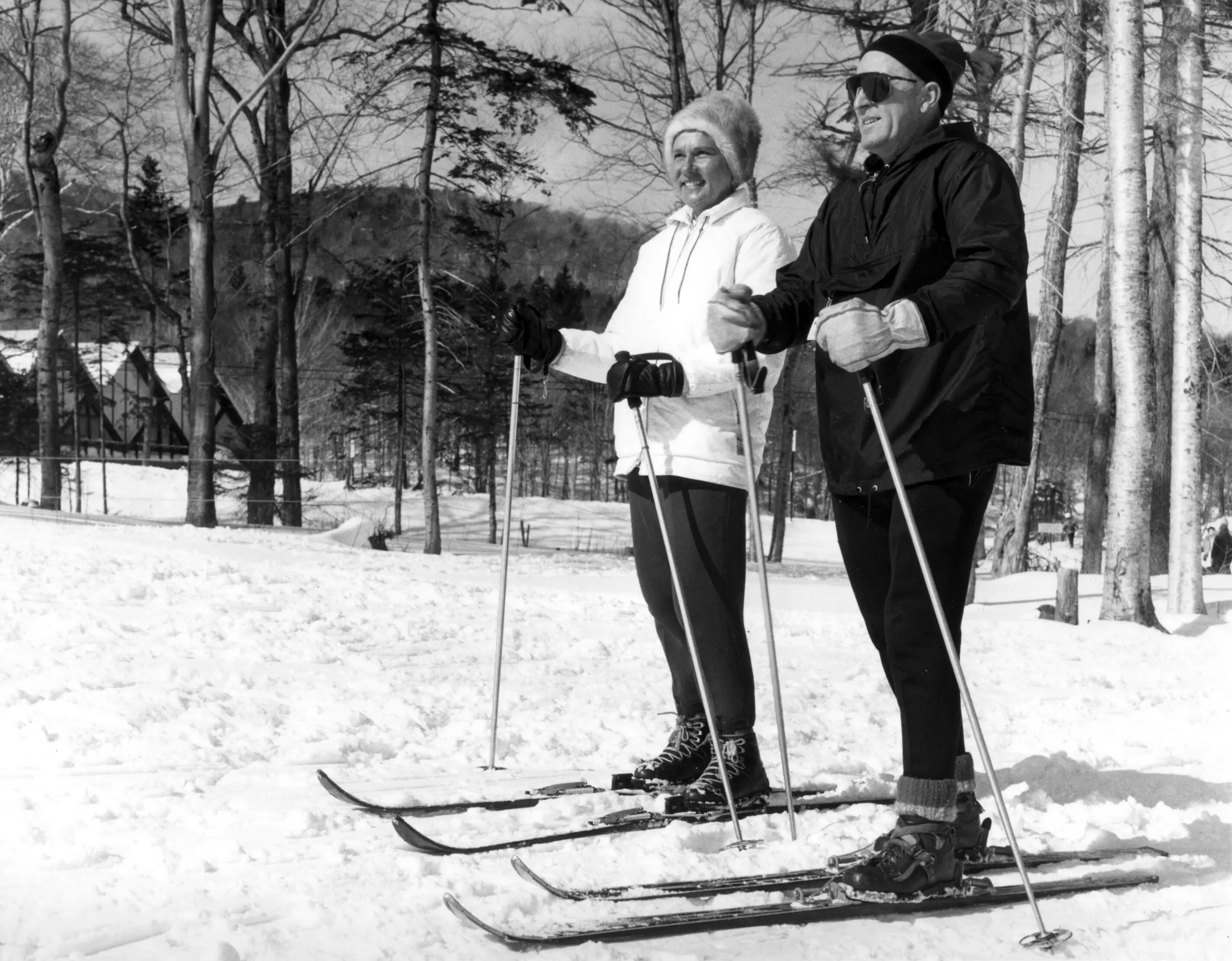 Two skiers pose on a snowy landscape, surrounded by trees in winter attire.