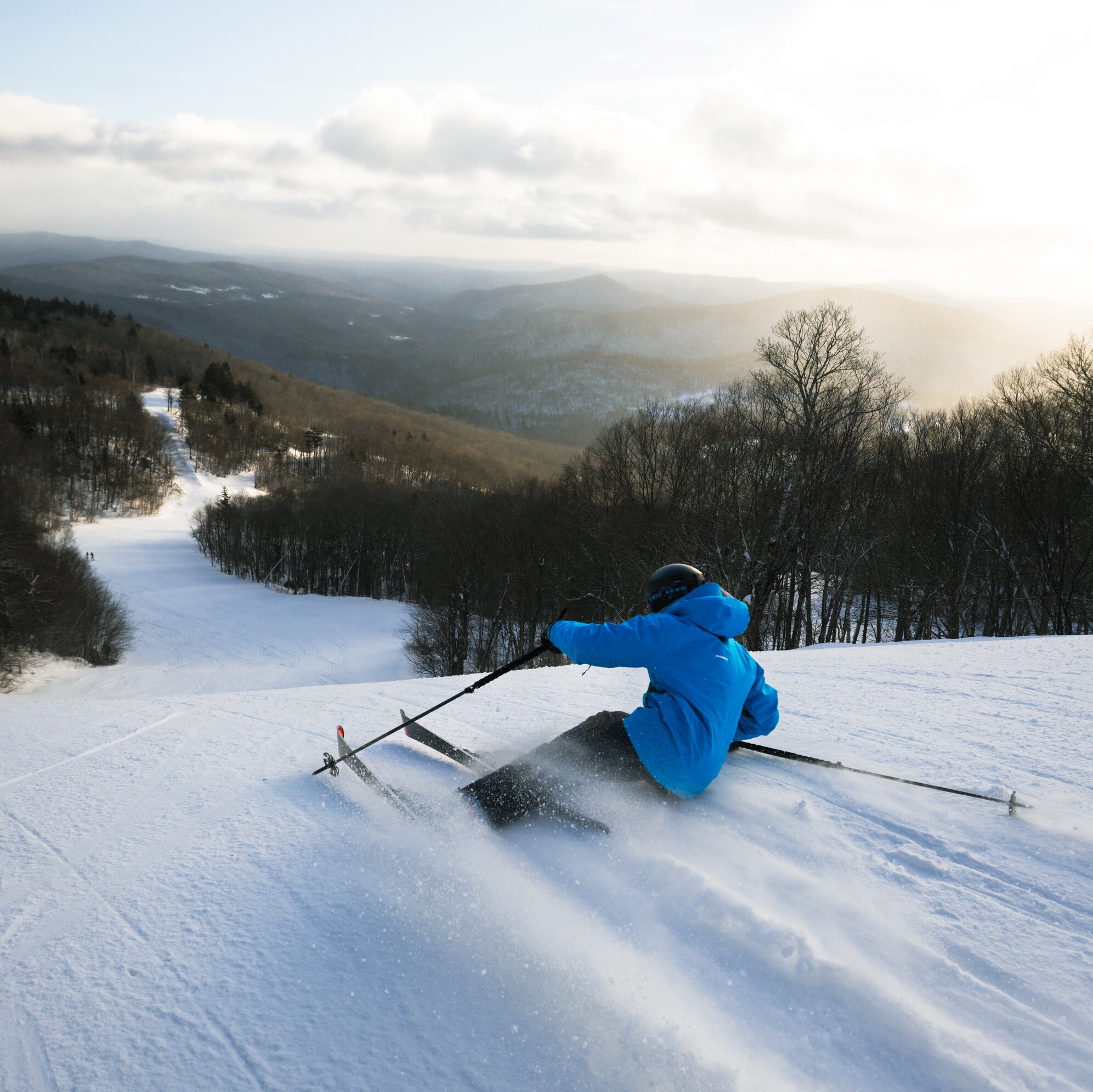 A skier in a blue jacket carves down a snowy slope with mountains in the background.