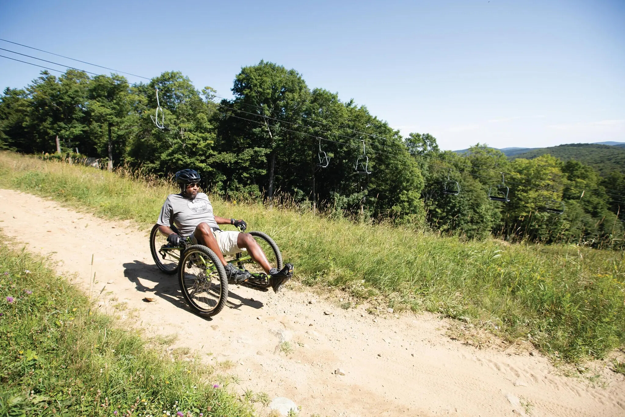 Person riding a handcycle on a dirt path surrounded by trees and grass.