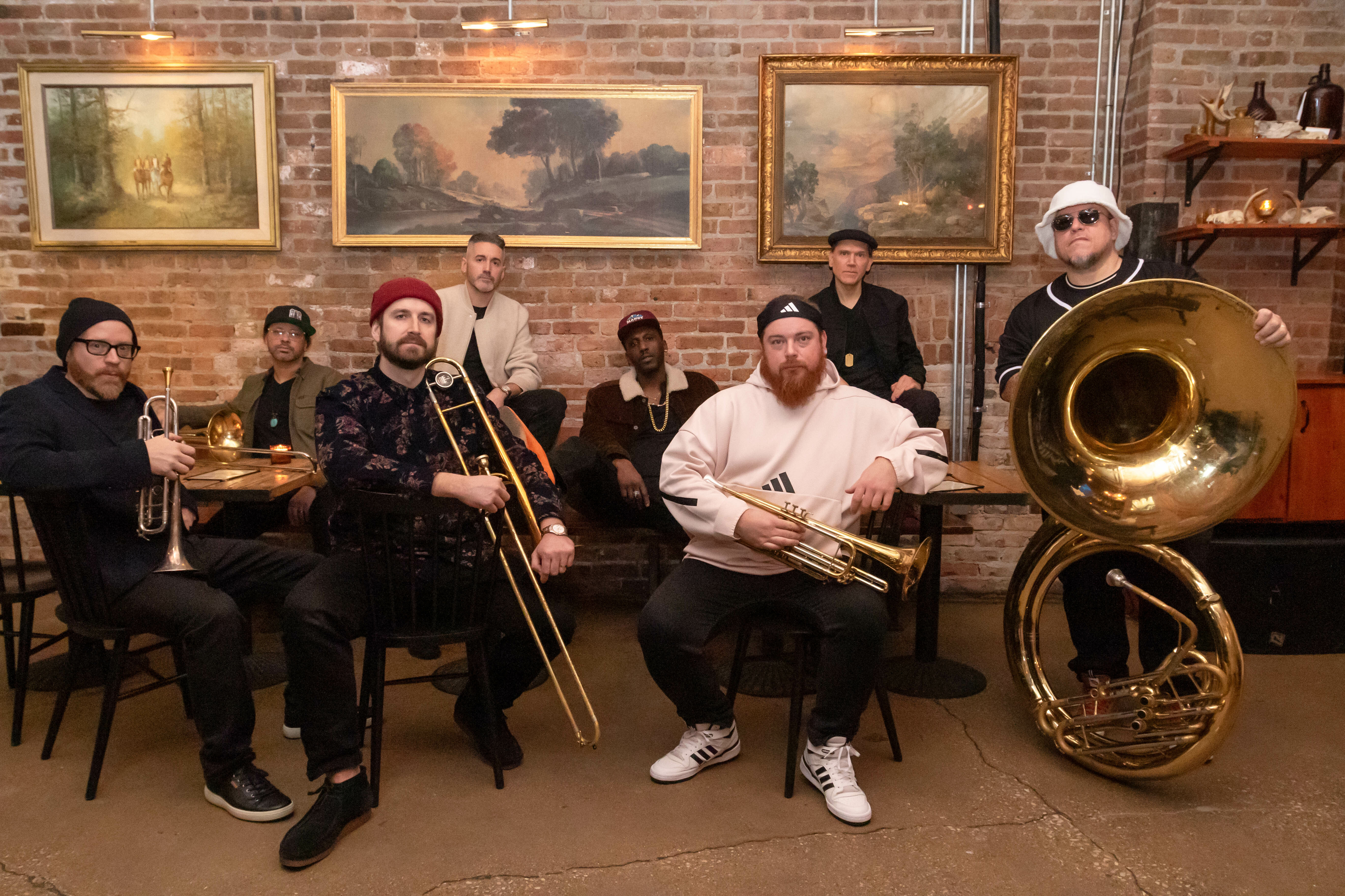 A group of eight musicians pose with brass instruments in a rustic setting.