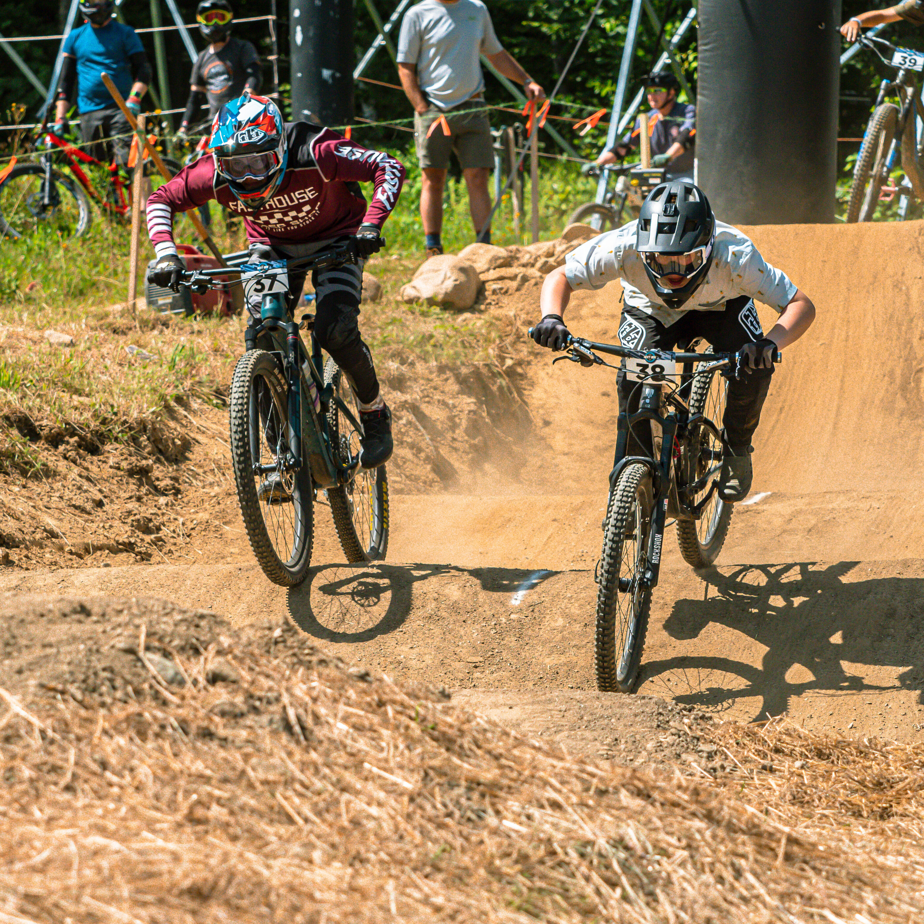 Two mountain bikers racing on a dirt track with spectators watching in the background.