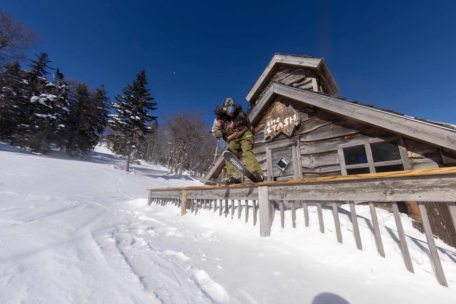 Snowboarder performing a trick on a railing near a wooden cabin in a snowy landscape.