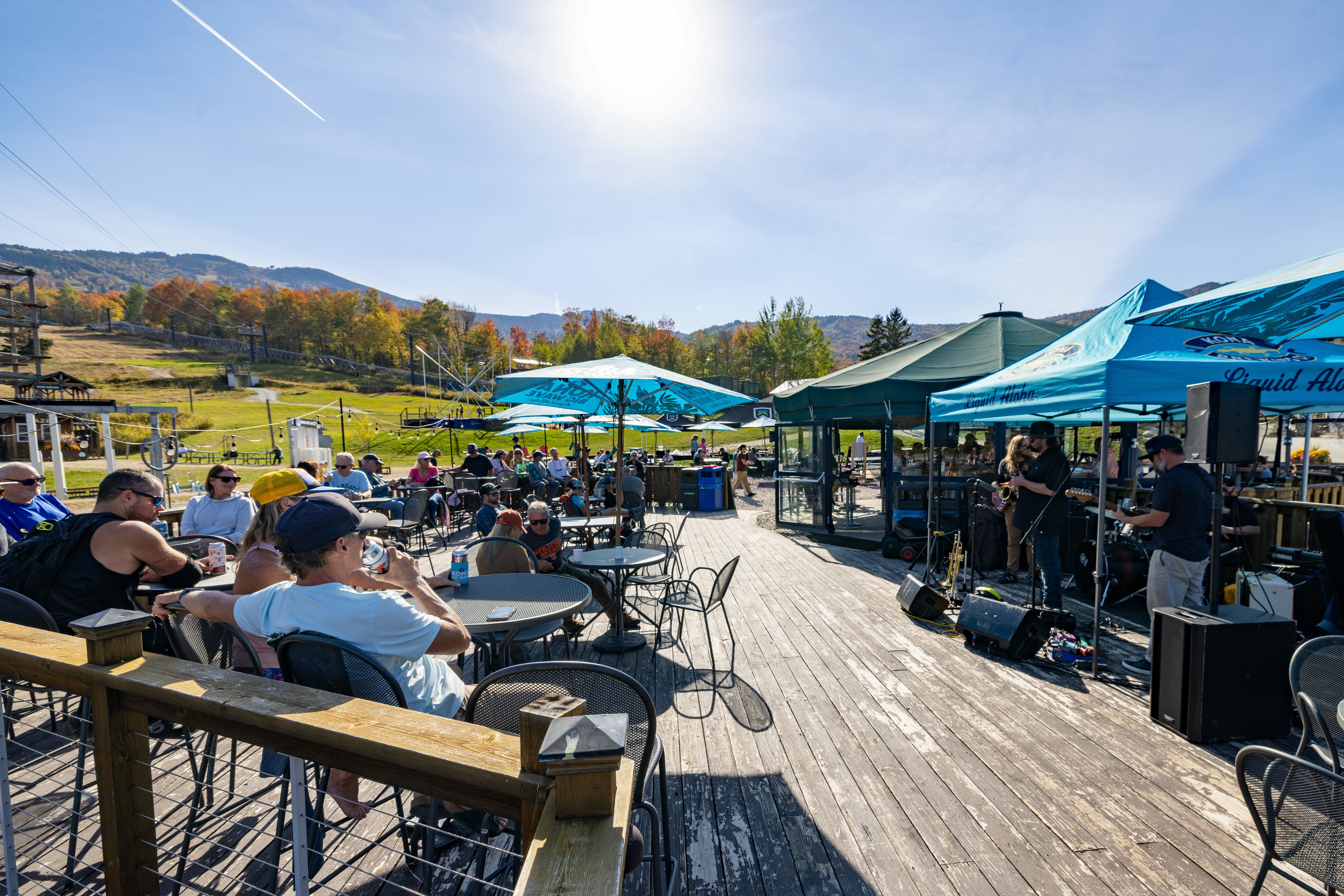 Outdoor event with people seated, enjoying music under umbrellas on a sunny day.