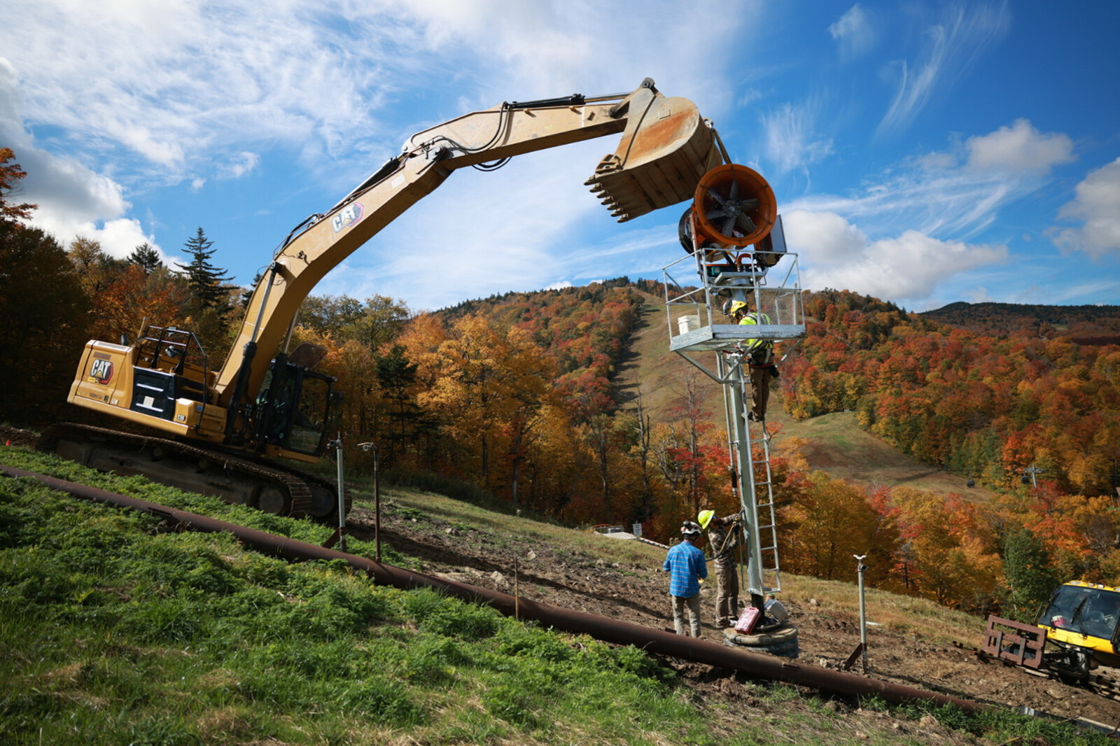 Excavator and crew working on a hillside with autumn trees in the background.