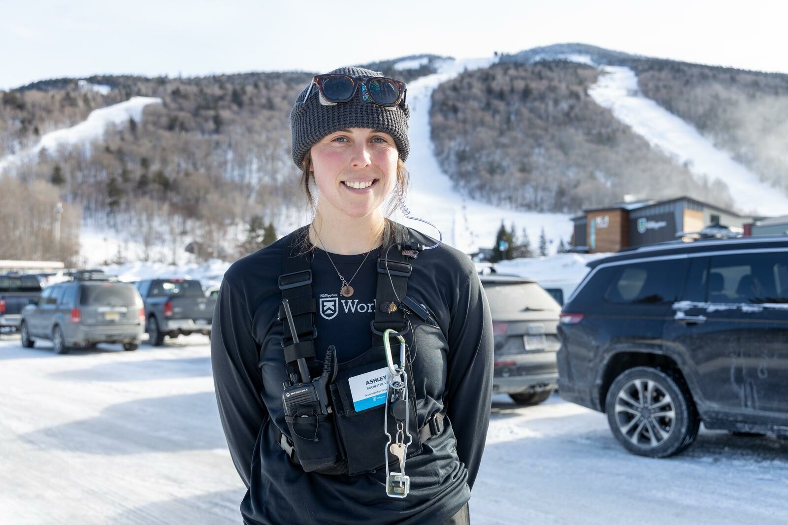 Smiling woman in winter gear stands in a snowy parking lot near ski slopes.