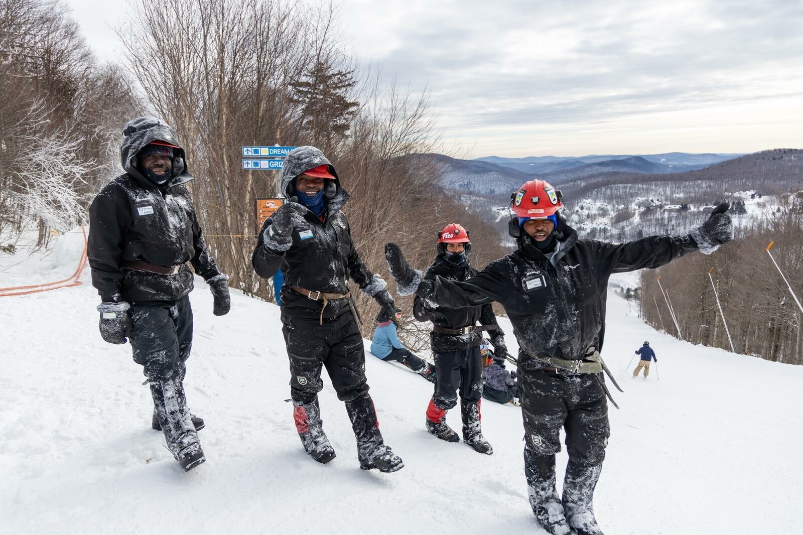 Four people in winter gear stand on a snowy slope, smiling and posing with ski signs in the background.
