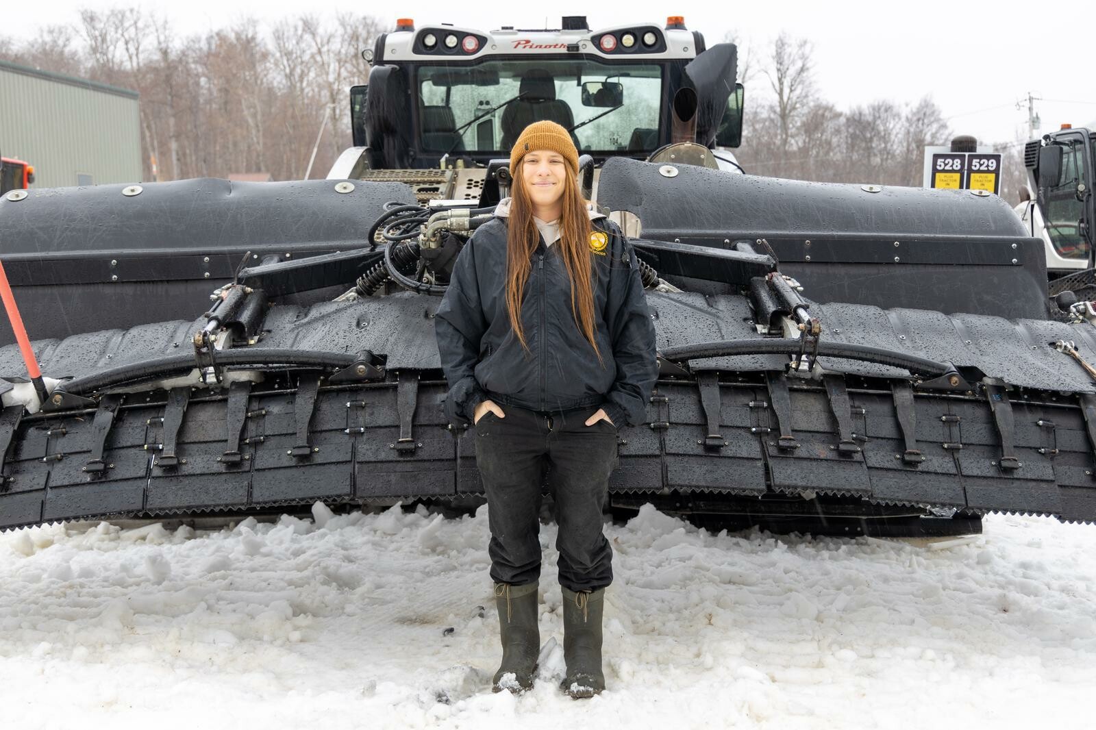 Person standing in snow with a large snowplow behind them on a winter day.