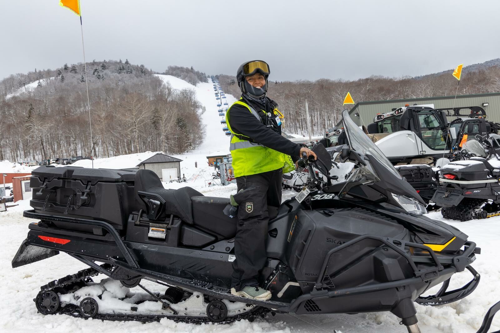 Person in a yellow vest smiles while sitting on a snowmobile in a snowy landscape.