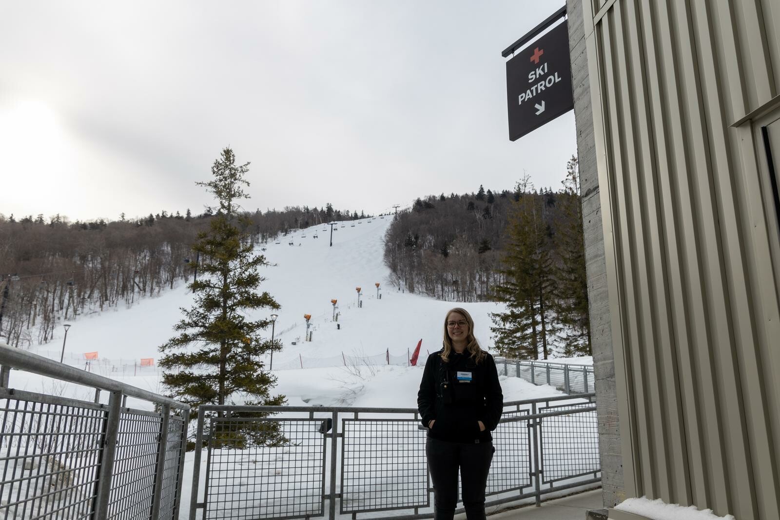 Ski patrol member stands at the base of a snowy ski slope with trees in the background.