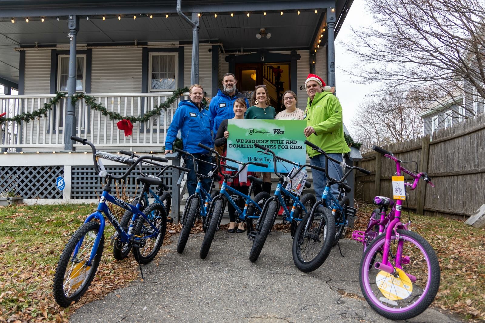 Group poses with donated bikes outside a decorated house, holding a sign for kids.
