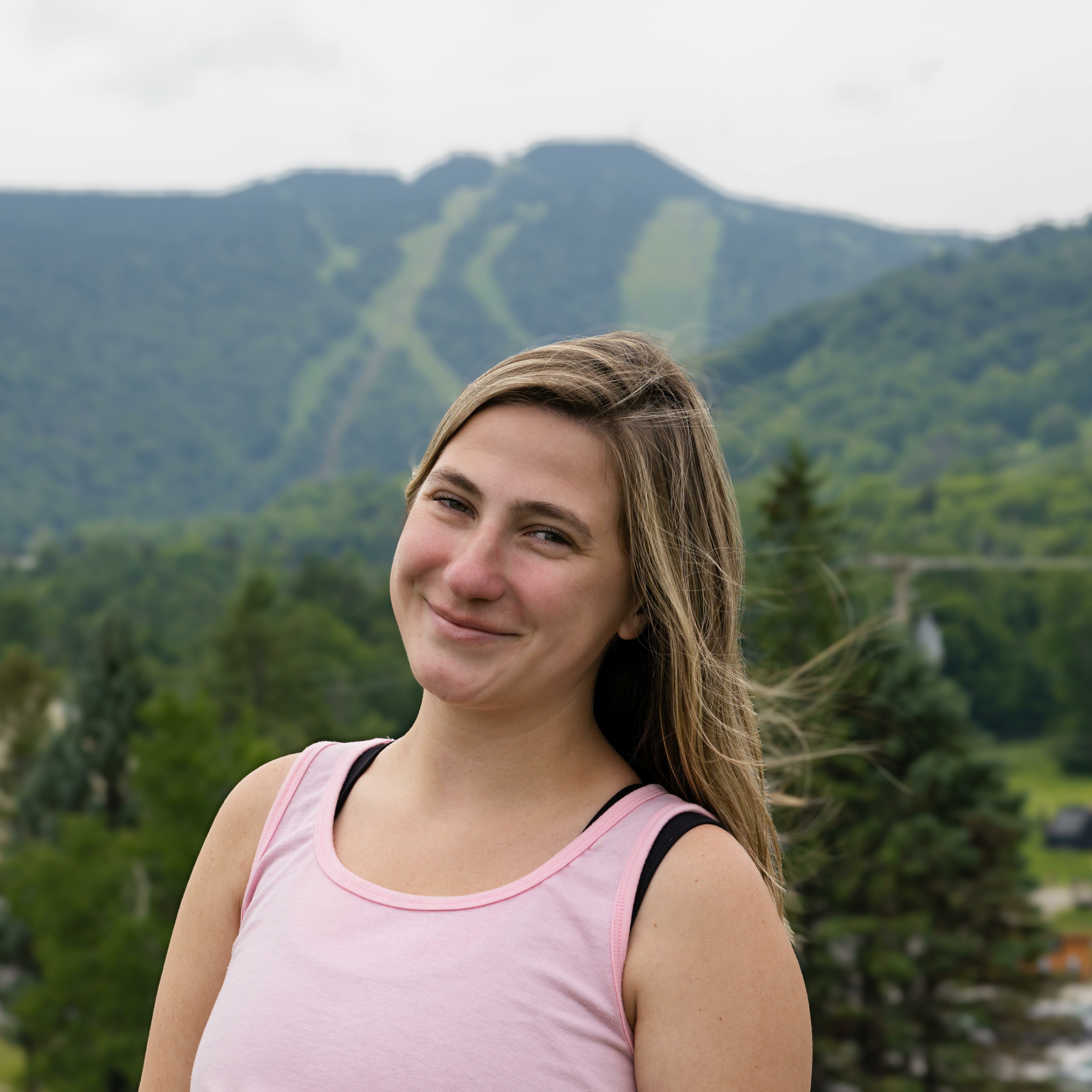 A smiling young woman in a pink tank top with mountains and trees in the background.