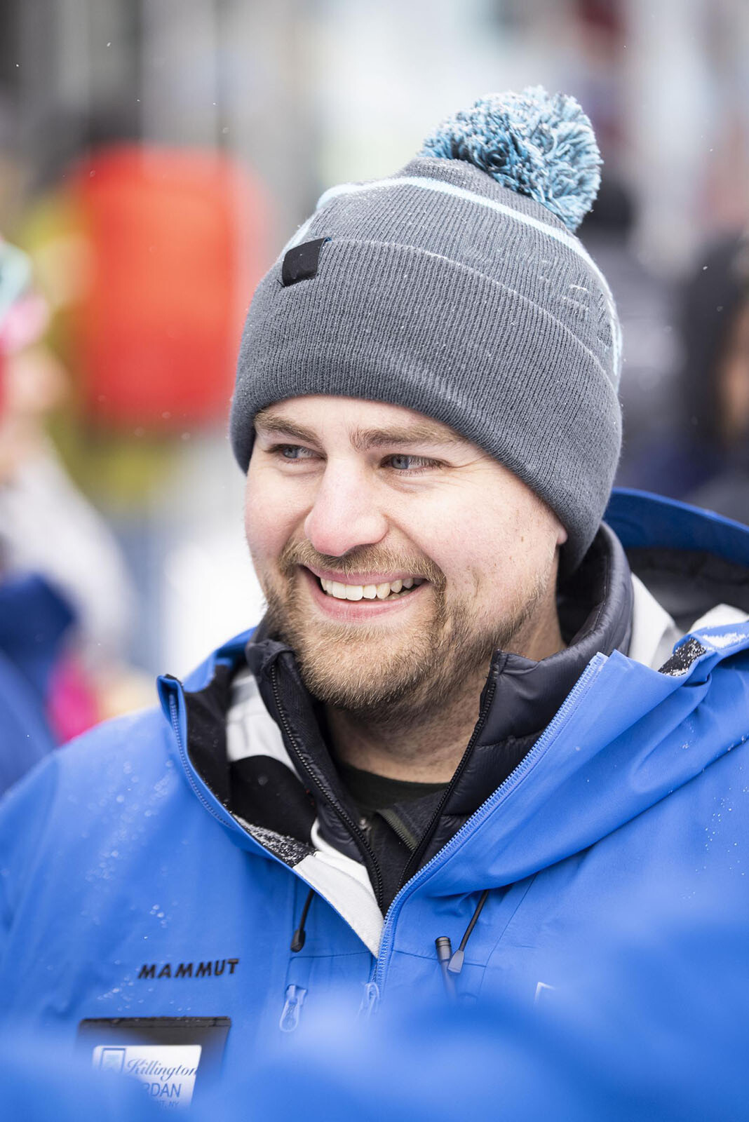 Smiling man in a gray beanie and blue jacket, outdoors in snowy surroundings.