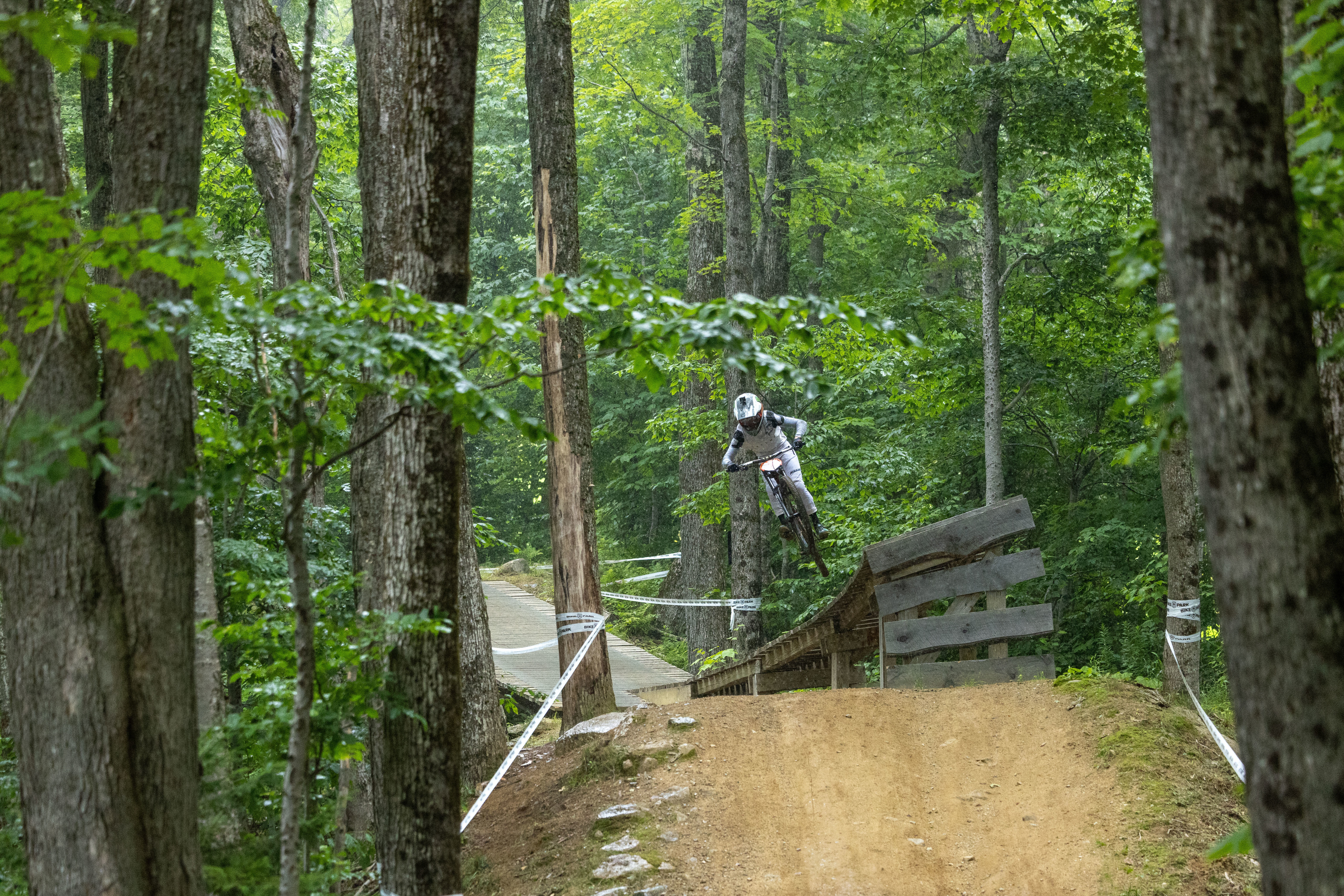 A mountain biker jumps off a wooden ramp in a lush green forest.
