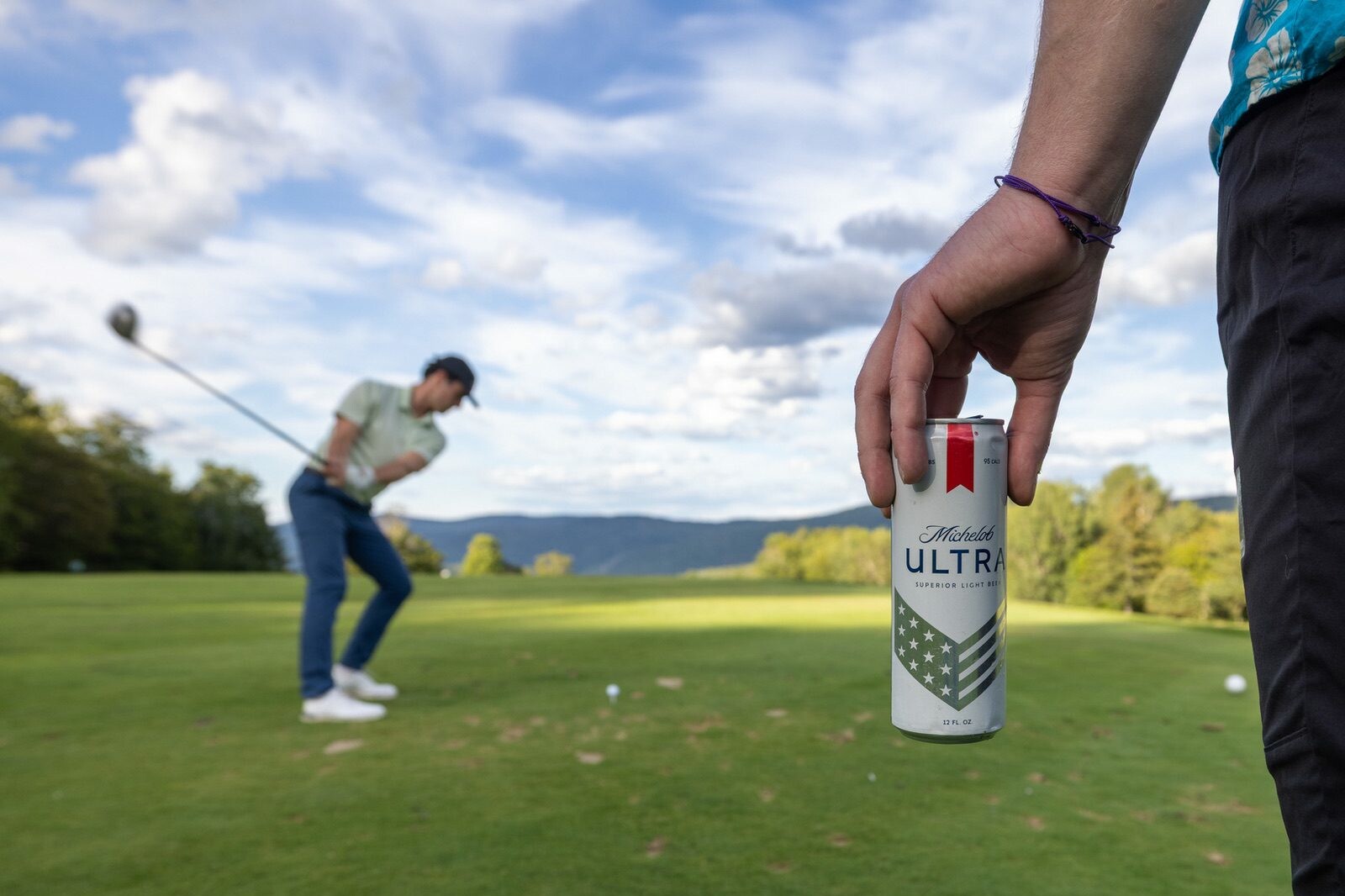 A golfer swings in the background while a hand holds a Michelob Ultra can in the foreground.